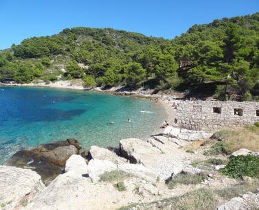 A secluded Croatian cove with clear turquoise water, a pebble beach, and lush green hills.