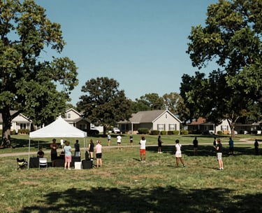 A wide-angle photography shot of a North American suburban community park where a wellness event is taking place. People are gathered under a white tent, with lush green trees and a clear blue sky. The scene is filled with a sense of stability and peace.