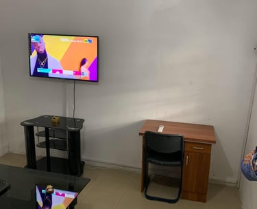 A living room with a wall-mounted TV over a glass stand, a wooden desk with a chair, and an AC unit.