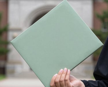 A close-up photograph of a graduate's hand holding a diploma folder, symbolizing academic success and opportunity, with a soft-focus North American university background in light gray and sage green tones.