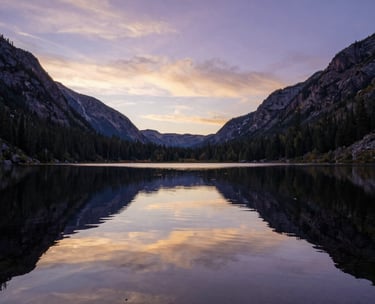 A wide-angle landscape photograph of a calm, reflective pond in a North American mountain valley. The water is still, mirroring a sky tinged with soft purple and gold at sunrise. The composition is peaceful and vast.