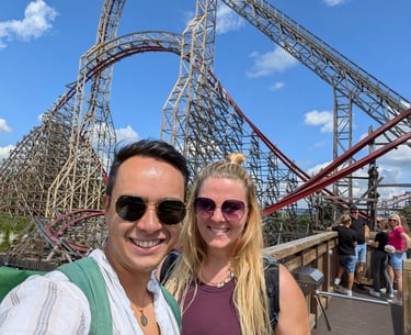 Two people smiling in front of a tall wooden rollercoaster on a sunny day with blue sky