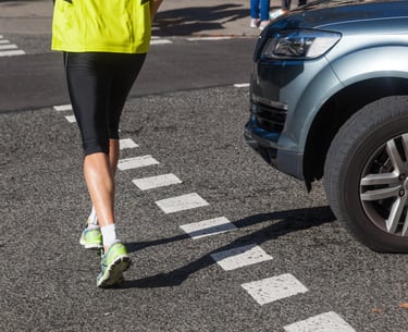 A runner in a neon yellow vest and leggings crossing a street in front of a blue car at a crosswalk.