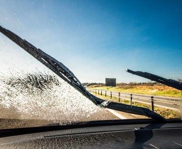 Car windshield wipers cleaning a dirty front window during a sunny drive on a highway.