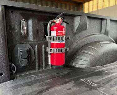 A red fire extinguisher mounted to the side wall of a pickup truck bed using a heavy-duty bracket.