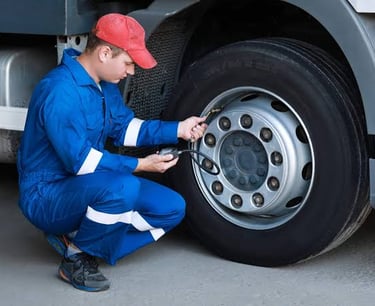 A mechanic in blue coveralls checks the tire pressure of a semi-truck wheel using a gauge.
