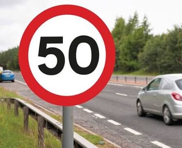 A circular red and white 50 mph speed limit sign on a roadside with cars passing by.