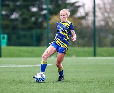 "Female soccer player in blue and yellow uniform dribbling on a field during a match.