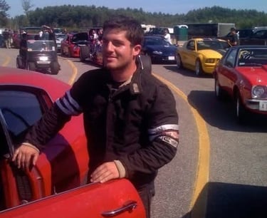 Don abenante racer in a black jacket stands by a red muscle car at a busy drag racing track.