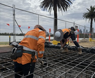 Construction workers in high-visibility safety gear install steel reinforcement mesh for a concrete foundation.