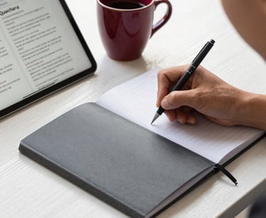 A close-up, high-quality photograph of a professional student's hands writing notes in an elegant charcoal grey notebook. Beside the notebook is a tablet displaying a quechua grammar lesson and a ceramic cup with deep crimson glazes. Soft, natural morning light on a white oak desk.