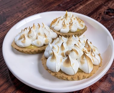 Three homemade mini lemon meringue tarts with toasted peaks on a white plate over dark wood.