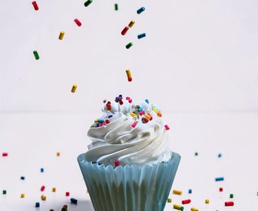 A vanilla cupcake with white swirl frosting being decorated with falling rainbow sprinkles.