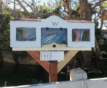 A white stucco Little Free Library shaped like a house with three glass window compartments filled with books.