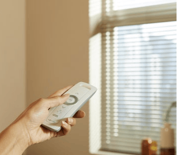 Hand using a remote control to operate motorized window blinds in a bright room.