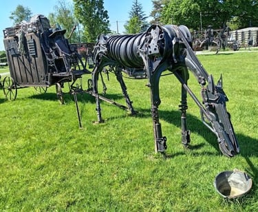 A life-sized steampunk horse sculpture made of scrap metal pulling a rustic carriage on green grass.
