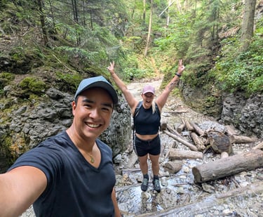 Two hikers smiling and posing on a rocky trail in a forest with fallen logs and greenery around