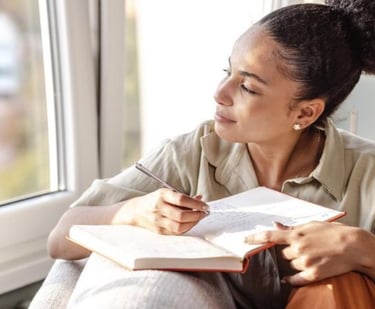 A young woman writing in her journal while looking out a window thoughtfully.