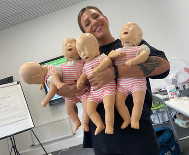 A person smiles while holding four infant CPR training manikins in a classroom setting.