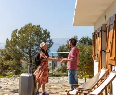 A woman with luggage checking into a sunny Mediterranean villa rental for a summer vacation.