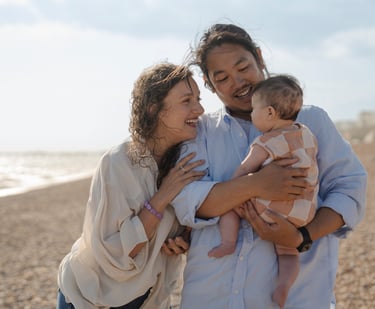 Family of three laughing on the beach with baby on the arms
