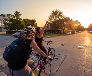 Image of two women bikers riding in the street, one with her arm stretched up in celebration