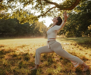 Eine Frau praktiziert Yoga im Krieger-1-Stand unter einem großen Baum in einer sonnigen Waldlichtung