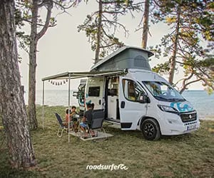 A family camping in a white pop-top camper van with an awning near a scenic ocean beach.