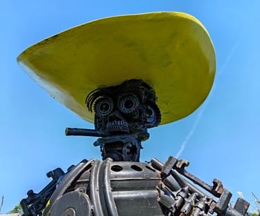 A metal scrap sculpture of a figure wearing a large yellow hat against a clear blue sky.