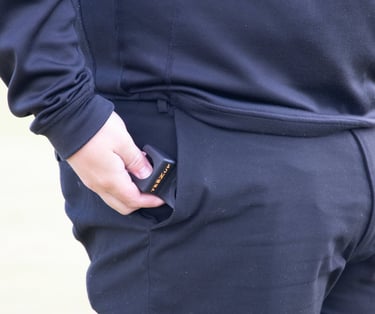 Golfer in black athletic wear placing a black and orange golf rangefinder into the right pocket of their trousers.