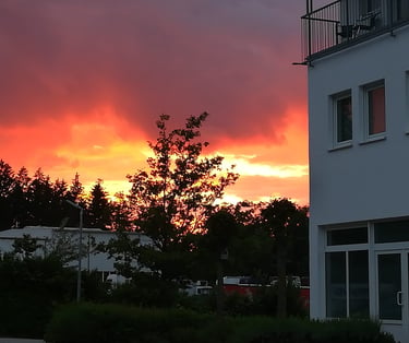 Vibrant orange and red sunset sky with dark clouds over a tree line and modern white hotel building.