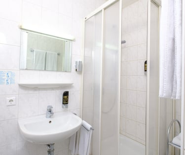 Compact hotel bathroom featuring a white pedestal sink, mirror, and a glass shower stall with white tile walls.