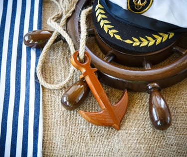A decorative captain's hat, placed on a ship's wheel with an ornamental wooden anchor next to it.
