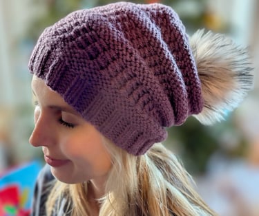 A young woman wearing a purple handknit slouchy beanie with a faux-fur pompom