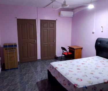 A studio with light purple walls, a bed with floral sheets, and a wooden study desk, fridge.