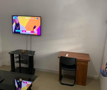 A living room with a wall-mounted TV over a glass stand, a wooden desk with a chair, and an AC unit.