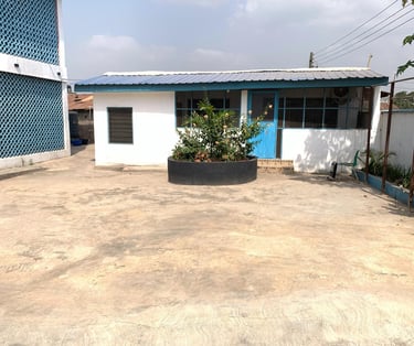 Small white building with blue doors and trim next to a courtyard with a circular stone planter.