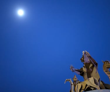 A metal bear sculpture silhouetted against a deep blue evening sky with the moon overhead.