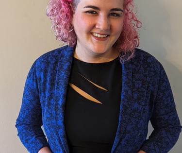 A woman with curly pink hair and an eyebrow ring, wearing a black dress and a blue blazer, smiles.