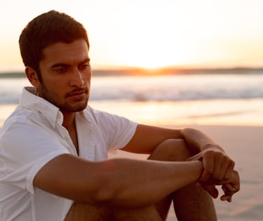 Hombre sentado en la playa al atardecer, con mirada reflexiva, simbolizando masculinidad consciente, conexión emocional y bie