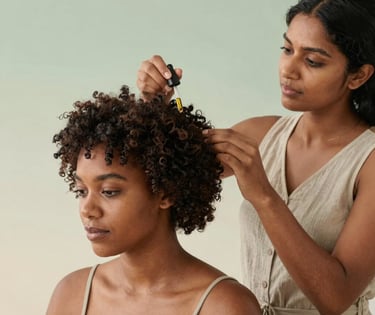 A woman applying essential hair oil treatment to another woman's curly hair with a dropper.