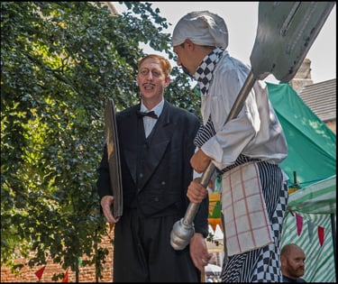 Street performers dressed as a comical chef and a formal butler act out a scene outdoors.