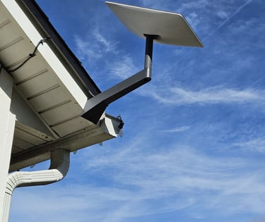 A Starlink satellite dish mounted on a residential roof eave against a clear blue sky.