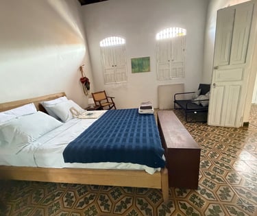 Rustic bedroom with blue bedspread, wooden bed frame, patterned tile floor, and white colonial shutters.