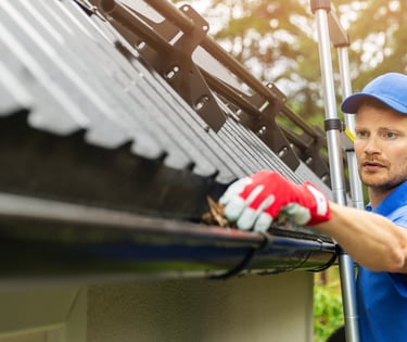 Professional contractor cleaning debris from a residential roof gutter system on a ladder.