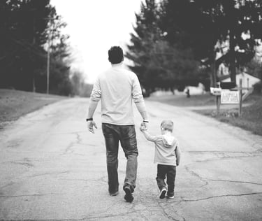 Black and white photo of a father and young son walking hand in hand down a quiet suburban street. 