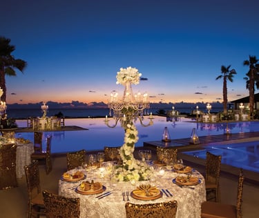 Luxury outdoor wedding reception by a pool at sunset with gold chairs and floral candelabra centerpieces.
