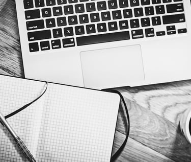Black and white flat lay of a laptop, graph paper notebook, pen, and coffee on a wooden desk.