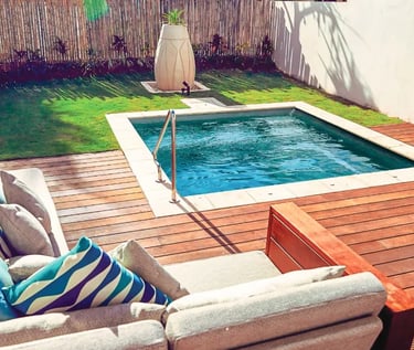 Compact square plunge pool with clear turquoise water and light stone coping around warm timber deck