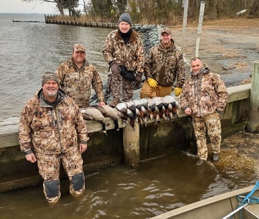 A hunting party posing on a dock in the water with a diverse harvest of ducks displayed in front of them.